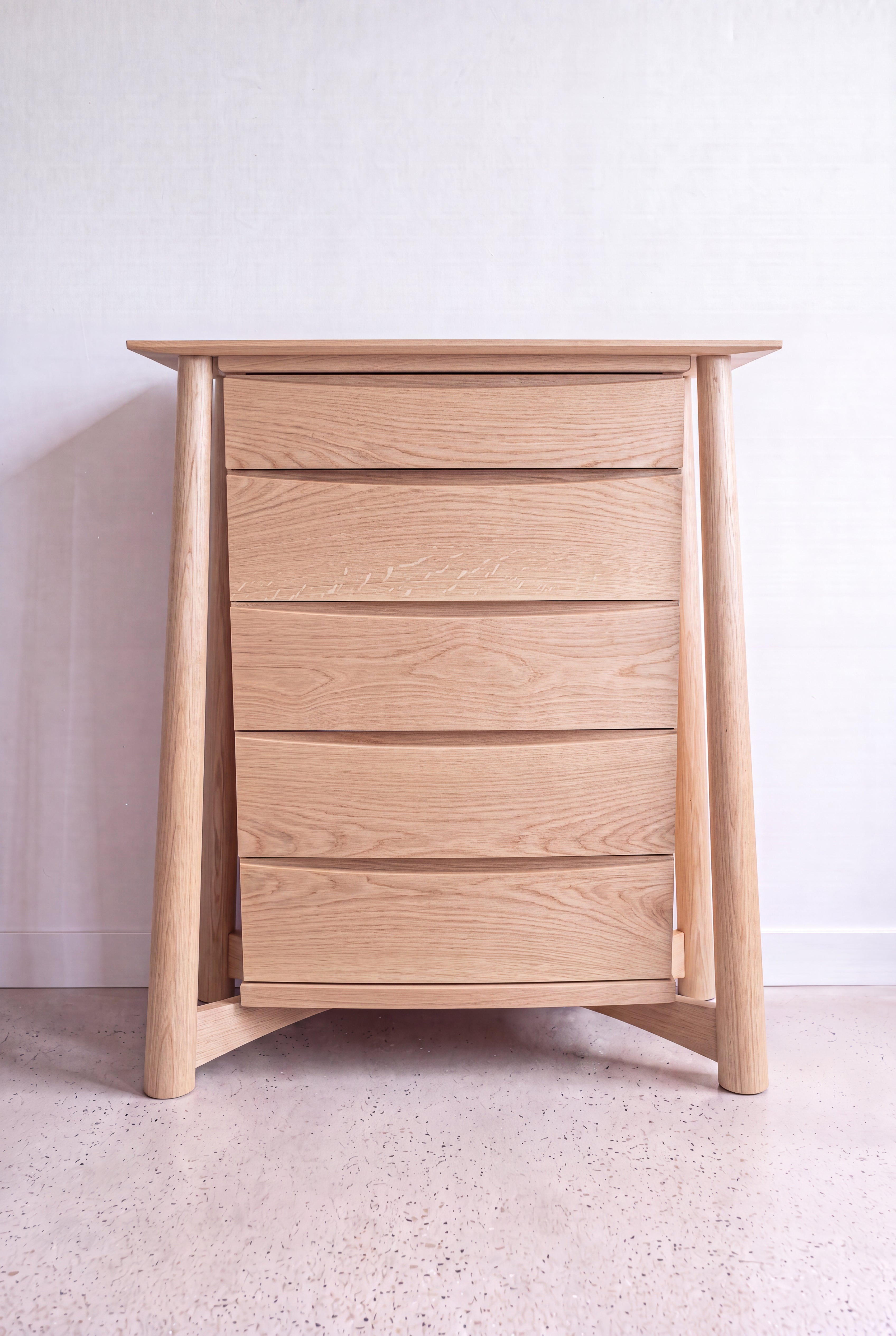 The Sunrise High Dresser by TY Fine Furniture, a handcrafted solid maple dresser with four drawers and angled legs, stands against a plain light wall on a speckled floor.