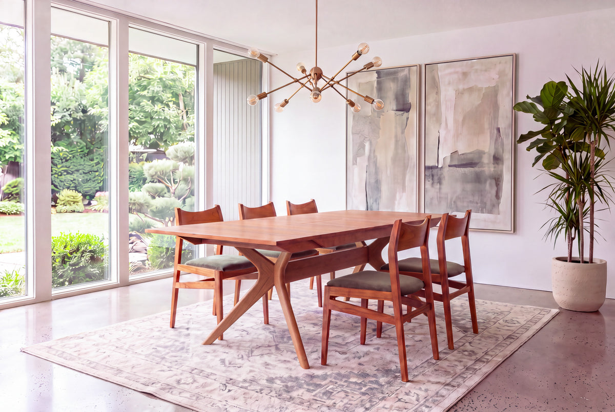 A modern dining room featuring the Solid Cherry Extendable Dining Table from the Strata Collection by TY Fine Furniture, surrounded by six chairs, with large windows offering views of a green garden, abstract art on the wall, a geometric chandelier overhead, and a tall potted plant in the corner.
