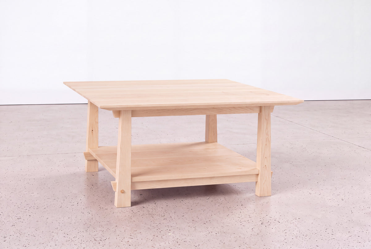 A square Solid Maple Horizon Coffee Table from the Horizon Collection by TY Fine Furniture, crafted in light wood with a lower shelf, sits on a speckled floor against a plain white wall. This elegant piece exemplifies the refined beauty and timeless craftsmanship of solid maple furniture by TY Fine Furniture.
