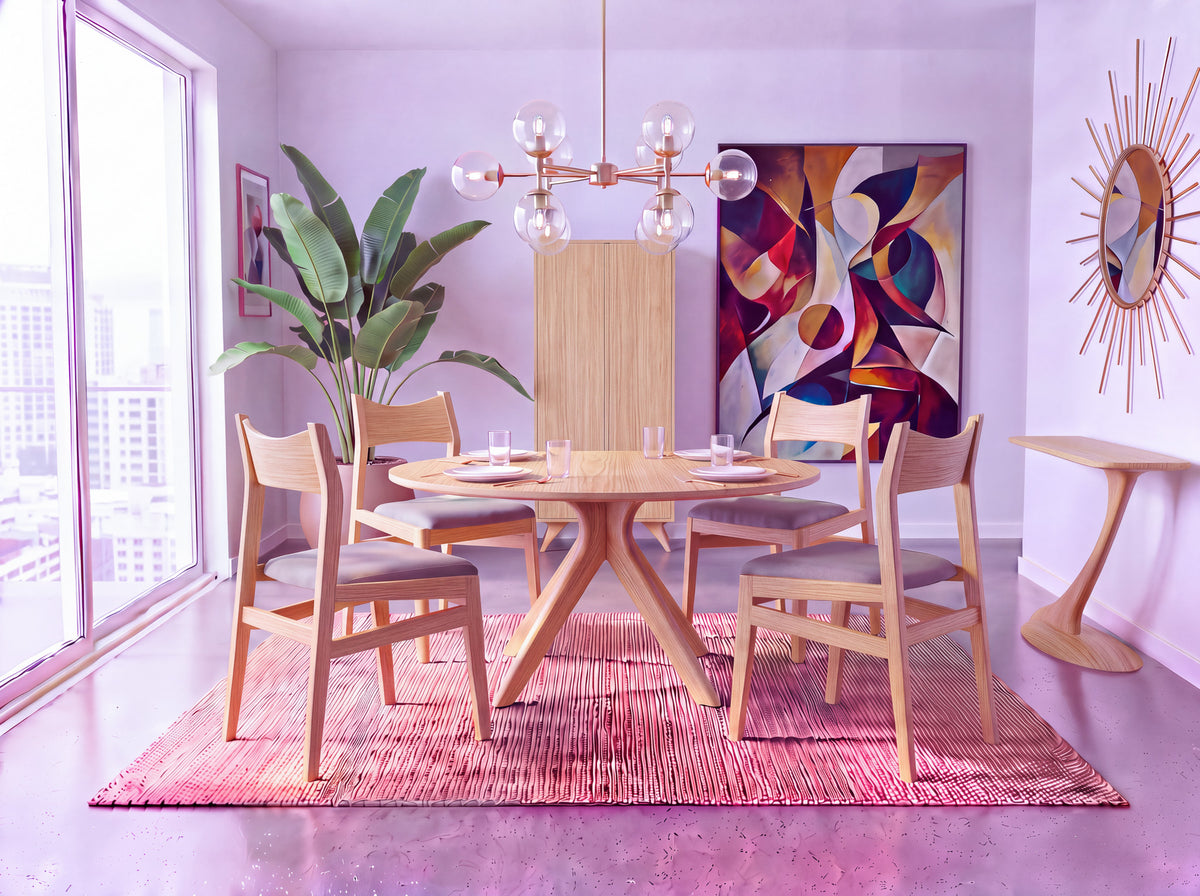 A modern dining room showcases the artisan-crafted Solid White Oak Round Dining Table from the Strata Collection by TY Fine Furniture, paired with four chairs. The space is accented by a pink rug, abstract wall art, a plant, a sunburst mirror, and a chandelier. Expansive windows reveal an impressive city view.