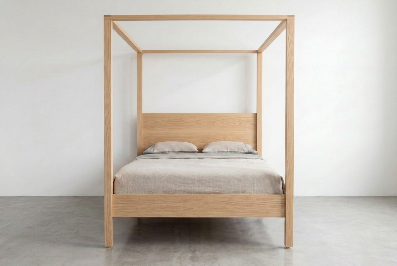 The TY Fine Furniture Modern Four Poster Bed—crafted from solid white oak—features a minimalist design with a simple headboard, two pillows, and a beige bedspread. Placed in a bright room with plain white walls and a concrete floor, this bed effortlessly blends clean lines with timeless materials for an understated and elegant look.