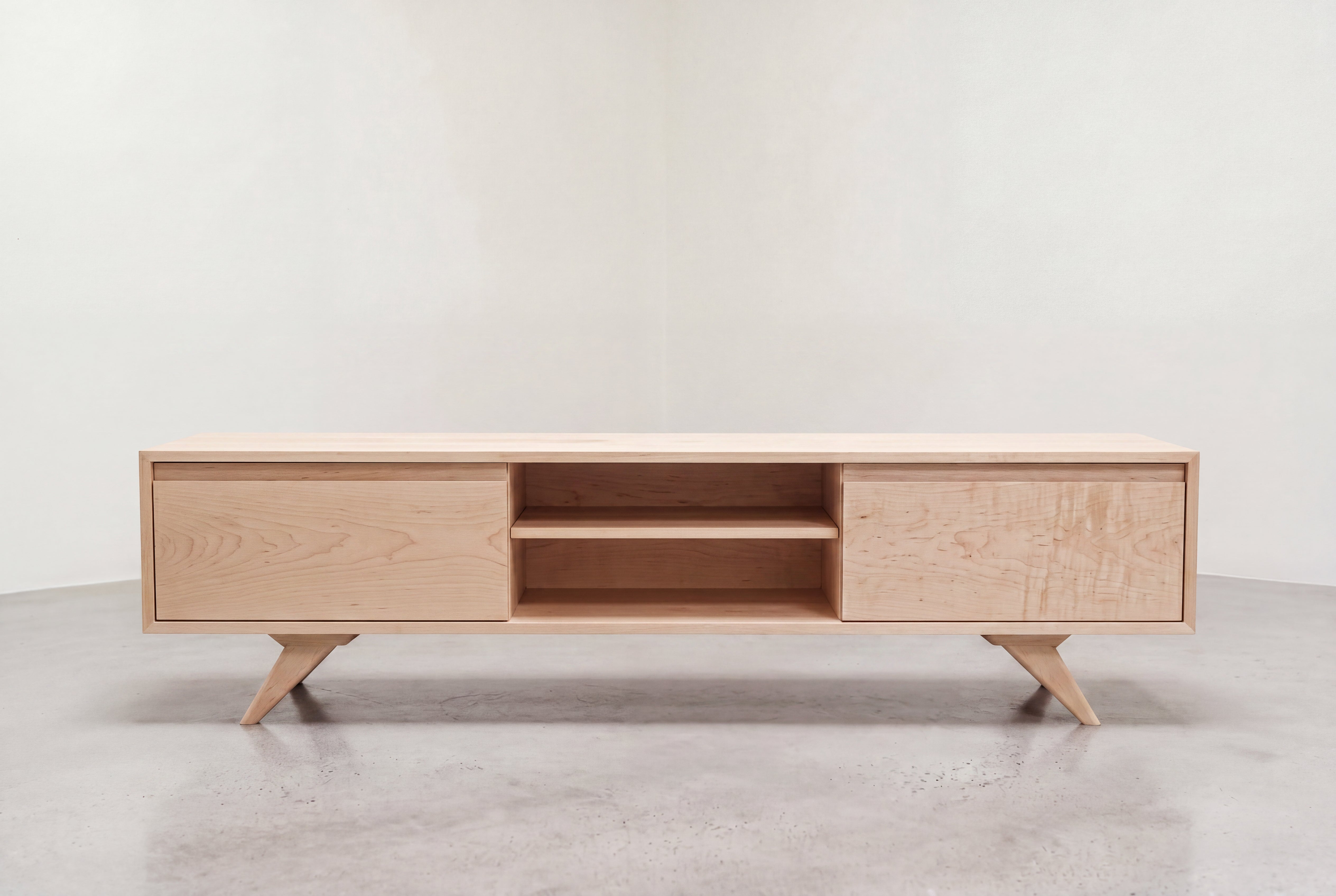 The Modern Entertainment Console with Drawers by TY Fine Furniture is a minimalist, modern TV stand crafted from solid wood. This piece features angled legs, two spacious drawers on each side for ample storage, and two open shelves in the center. It is shown resting on a smooth concrete floor against a plain white wall.
