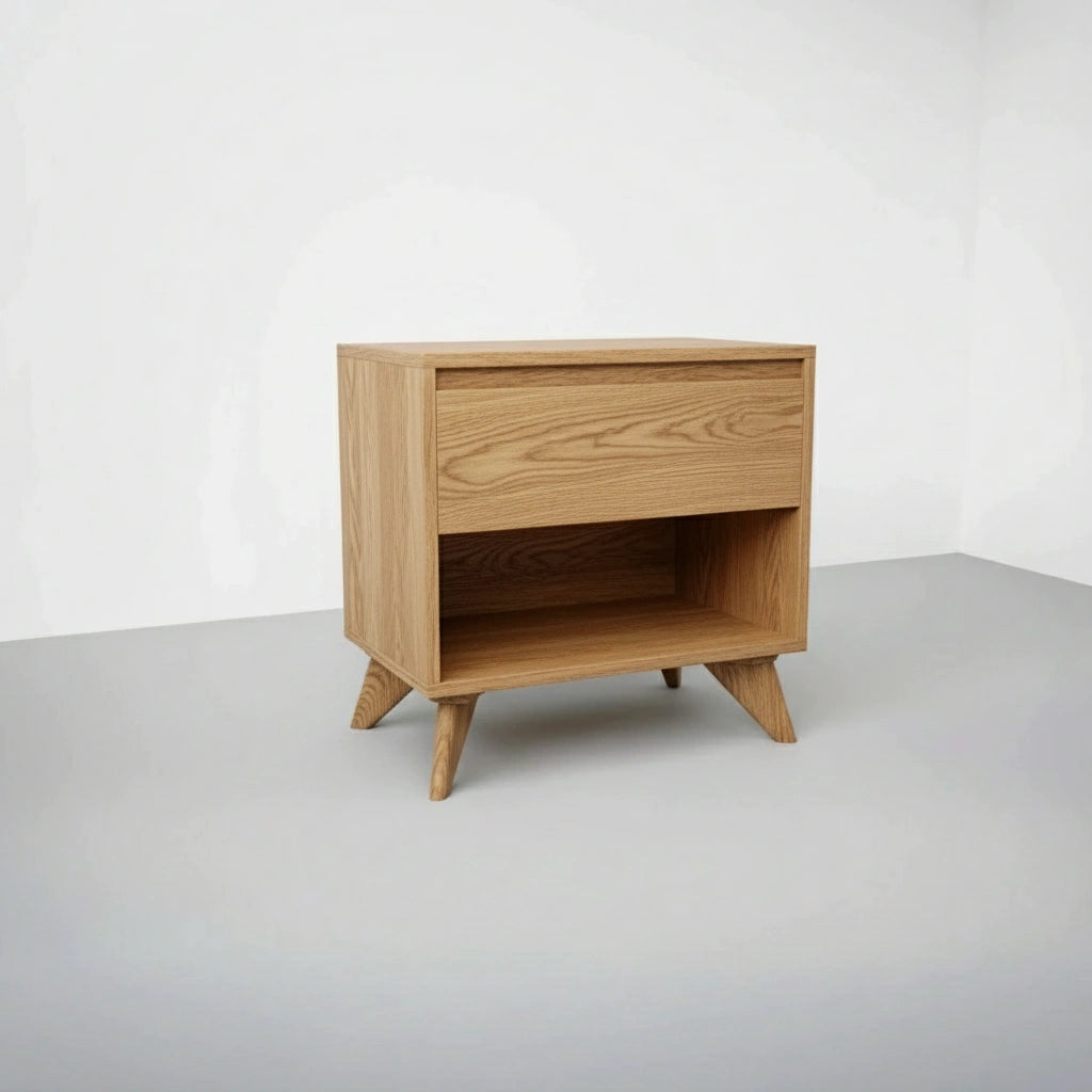 The Modern Cube Side Table - White Oak by TY Fine Furniture is crafted from solid wood with angled legs, a push-to-open drawer, and an open shelf, styled against a plain white wall and gray floor.