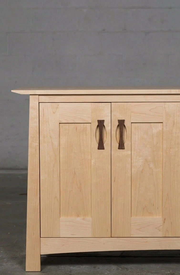 The Enso Solid Wood Credenza Buffet by TY Fine Furniture features handmade maple, two panel doors with dark curved handles, and stands on a concrete floor against a gray brick wall—ideal sustainable storage for modern dining rooms.