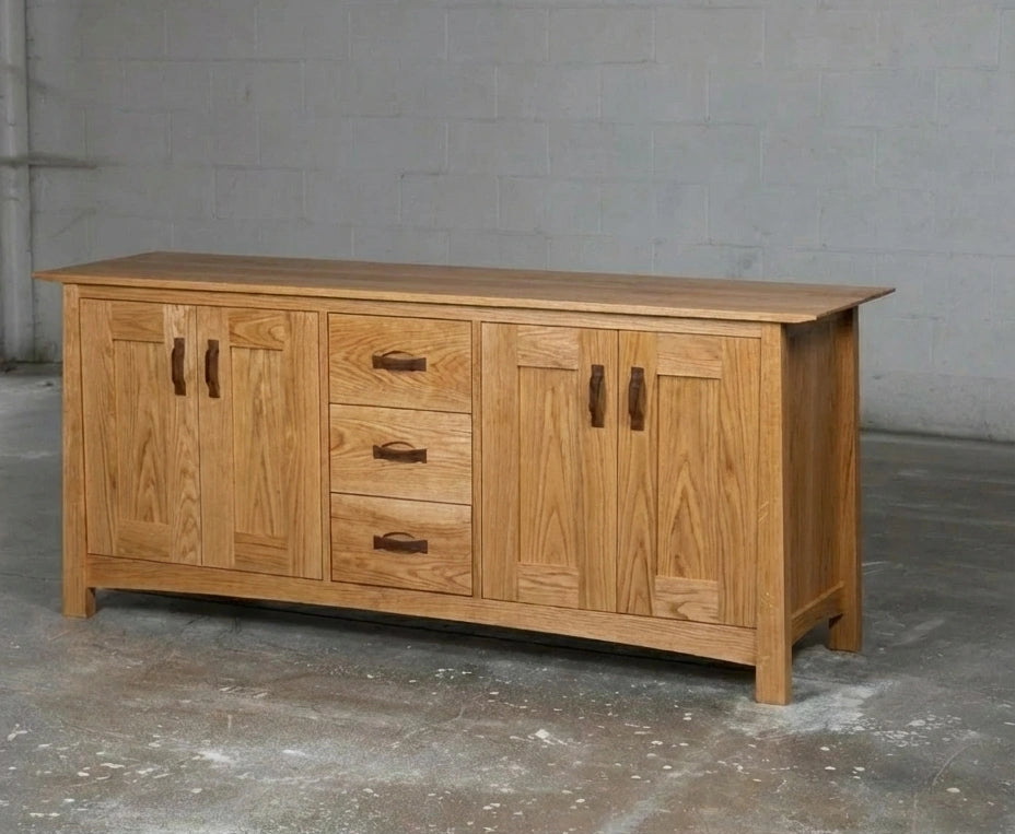 The Enso Solid Wood Credenza Buffet by TY Fine Furniture, handmade in white oak, features dual side cabinets and three central drawers for stylish dining room storage, shown on a concrete floor against a plain gray wall.