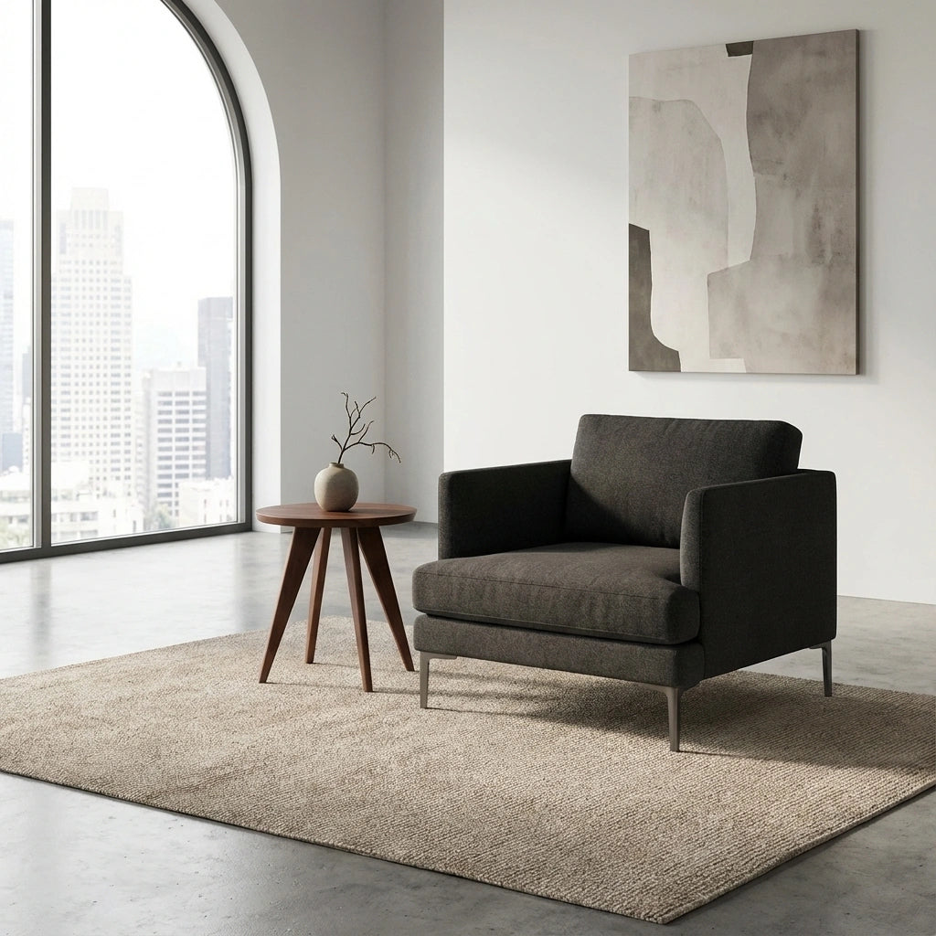 A modern living room features a dark gray armchair, a TY Fine Furniture Essentials Solid Wood Round Side Table—handcrafted in walnut, a minimalist vase, beige rug, abstract wall art, and a large window with city views.