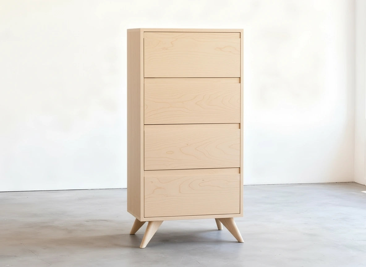 The Tall Modern Dresser Mid Century in maple by TY Fine Furniture, featuring four drawers and angled legs, stands on a smooth gray floor against a white wall in a minimalist room.