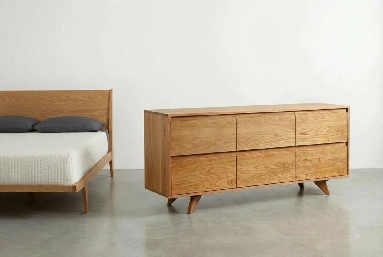 A minimalist bedroom features a wooden bed with gray pillows, white blanket, and the TY Fine Furniture Modern Dresser in solid white oak, both set on a light gray floor against a plain white wall.